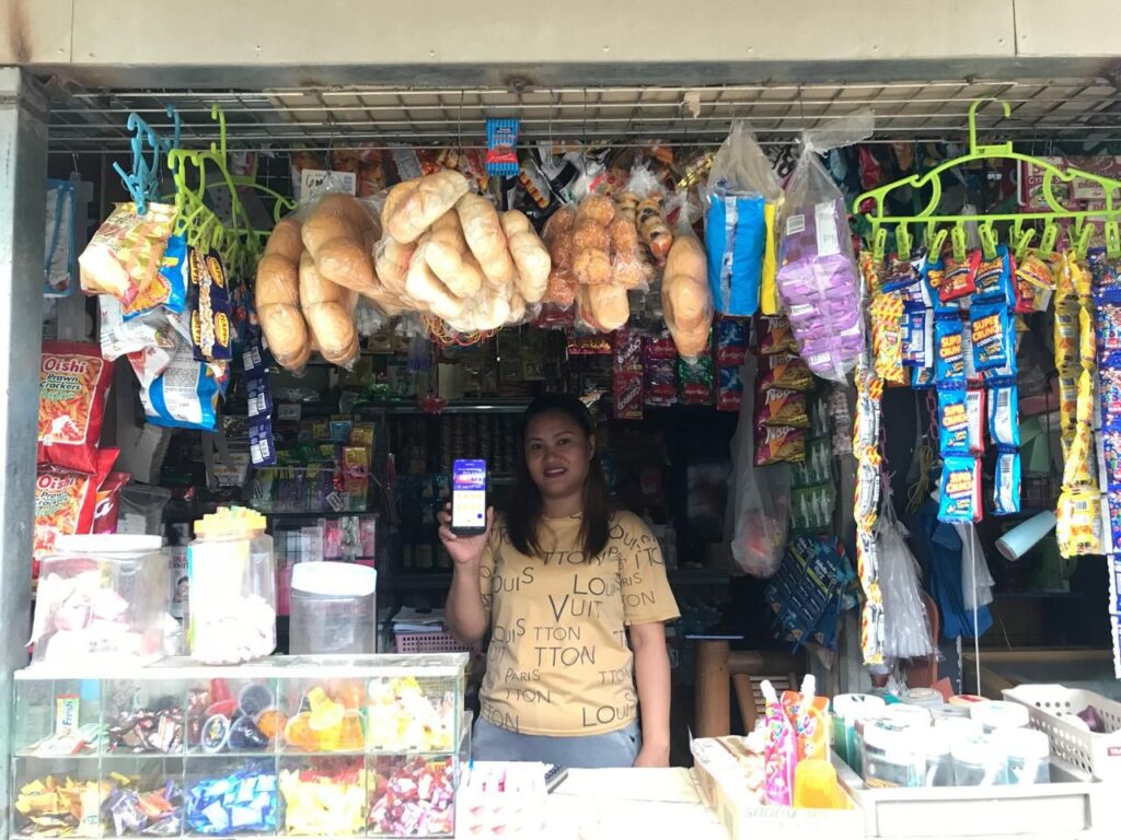 A yufin user behind the counter of her sari-sari store, holding up her smartphone displaying the yufin app.