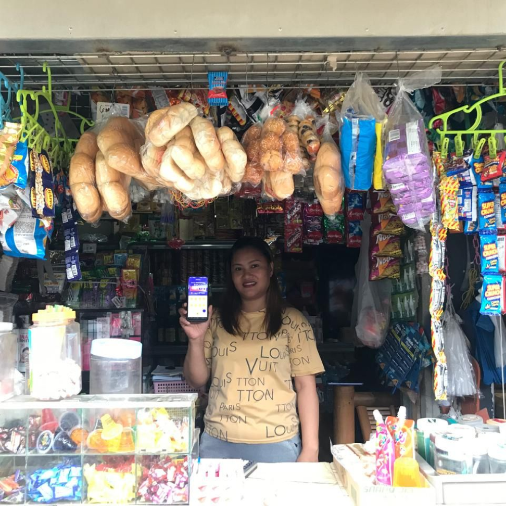 A yufin user behind the counter of her sari-sari store, holding up her smartphone displaying the yufin app.