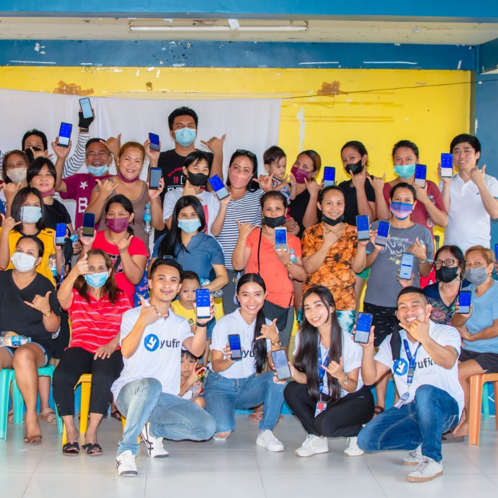 The yufin team with a group of people holding up their smartphones displaying the yufin app, while forming the yufin symbol with their hands.